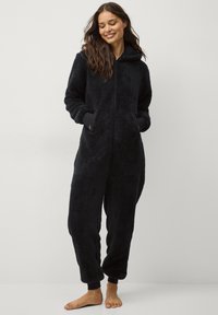 Zwarte fleece onesie met ritssluiting aan de voorkant, capuchon en lange mouwen. Bevat zijzakken en aansluitende manchetten op de benen. Zachte, gestructureerde stof.