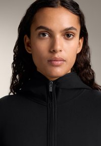 Črna zip-up kapuca s gladko teksturo, ki ima osrednji zadrgo in prilegajoč obliko. Preprost in minimalističen slog.