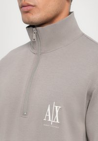 Sudadera gris claro con cuello alto y cierre de cremallera, que presenta un logotipo blanco de Armani Exchange en el lado izquierdo del pecho. Textura suave.