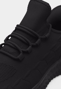 Μαύρο αθλητικό παπούτσι με πλεκτό ύφασμα επάνω μέρος, ελαστικούς βρόχους κορδονιών και μαξιλαρωμένη, υφή σόλα, παρουσιάζεται σε κοντινή λήψη.