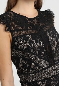 Robe en dentelle noire avec des motifs floraux complexes, coupe ajustée, et des accents doux de volants sur les épaules, présentant des sections transparentes et opaques.