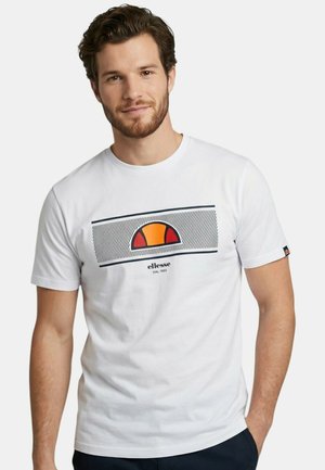 Jeune homme aux cheveux bruns et à la barbe, portant un t-shirt blanc Ellessee avec un logo orange et rouge, debout avec une main dans la poche.