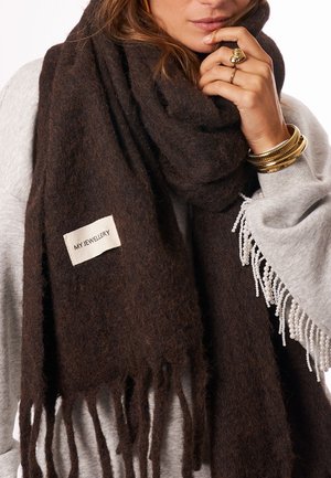 Vrouw met een dikke, donkerbruine franjesjaal met het label "MY JEWELLERY", een grijze trui met parelrand, en gouden ringen en armbanden.