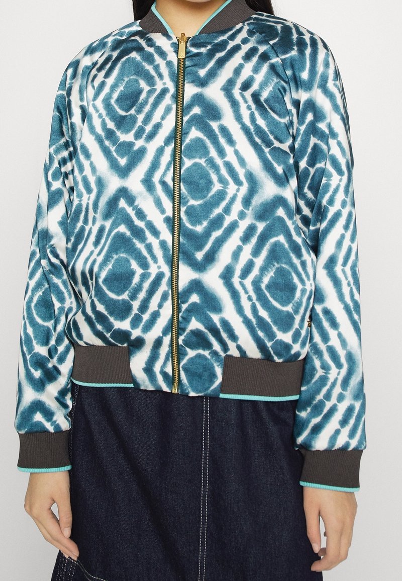 Lehká bomber bunda s modro-bílým vzorem tie-dye, zlatým zipem, hnědými žebrovanými manžetami a lemem s tyrkysovým akcentem.