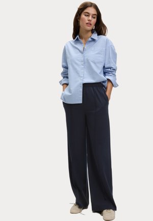Camicetta azzurro chiaro con maniche arrotolate e taschino sul petto, abbinata a pantaloni navy a vestibilità ampia e scarpe neutre. Design e tessuto semplici.