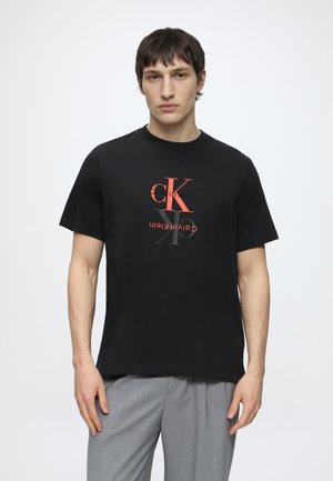 Muž s krátkymi tmavými vlasmi nosiaci čierne tričko Calvin Klein s červeným a sivým logom, v kombinácii so svetlosivými nohavicami, stojaci na bielom pozadí.