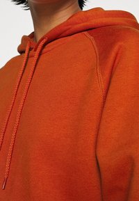 Sudadera naranja hecha de tela suave, con un bolsillo canguro, capucha con cordón y costuras texturizadas a lo largo de las costuras.