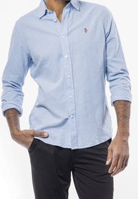 Camisa de botón de color azul claro en una tela texturada con mangas largas, que presenta un pequeño diseño bordado en el pecho, combinada con pantalones negros.