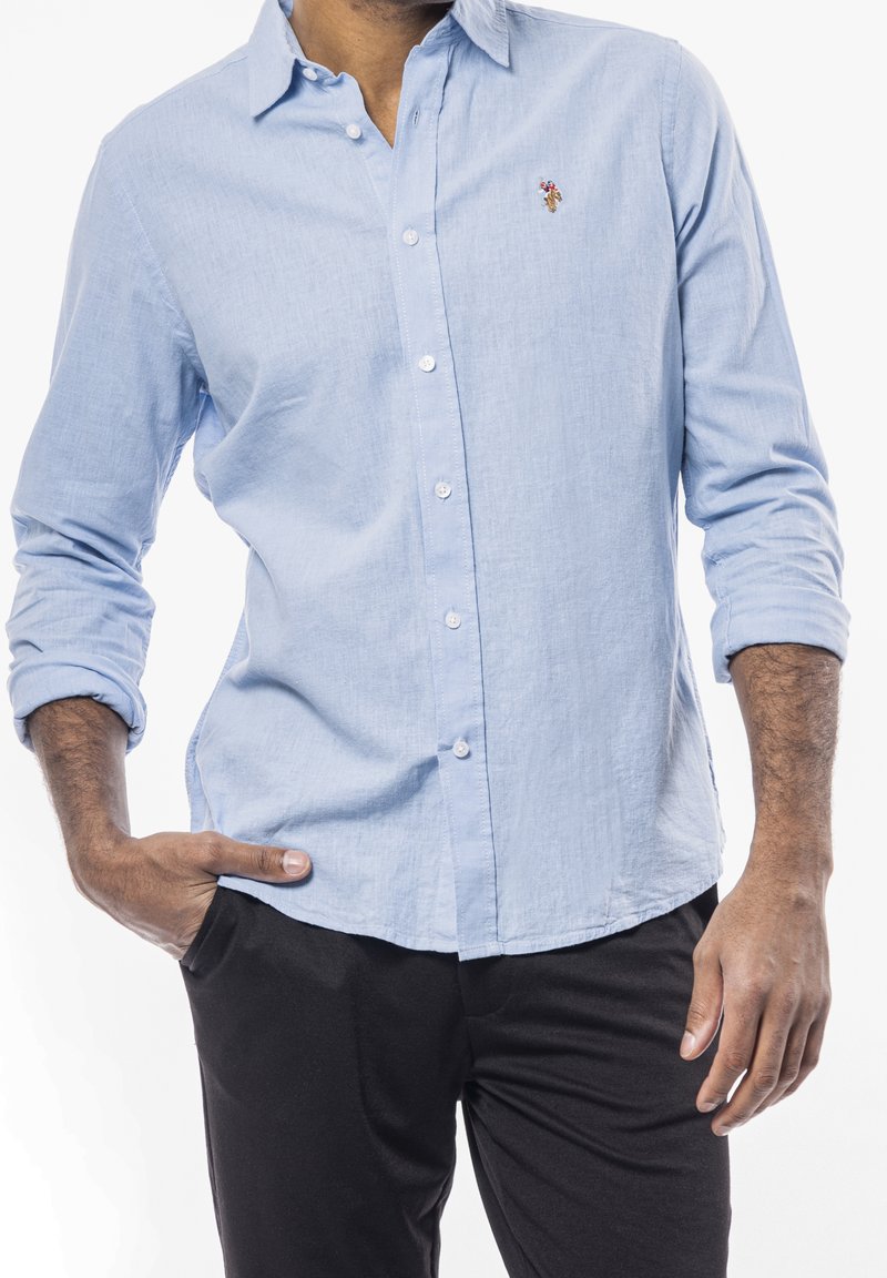 Camisa de botón de color azul claro en una tela texturada con mangas largas, que presenta un pequeño diseño bordado en el pecho, combinada con pantalones negros.