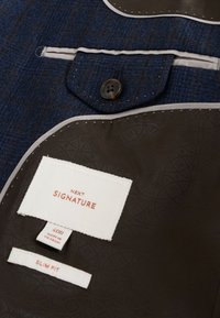 Veste texturée bleu foncé avec détail de boutons et étiquettes cousues indiquant Next Signature, 40R Made in Vietnam et Slim Fit sur une doublure marron à motif.