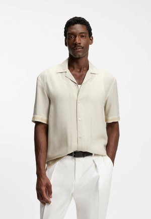 Mand iført kortærmet beige skjorte med subtilt mønster, hvide bukser og sort bælte, stående foran en ensartet hvid baggrund.