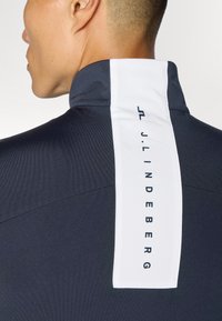 Dark navy athletic jacket with a stand-up collar; features a white logo patch at the back with text and subtle textured fabric.