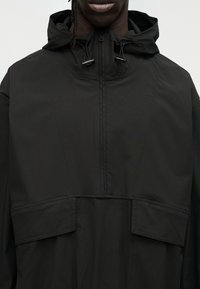 Μαύρο μπουφάν με κουκούλα κατασκευασμένο από απαλό ύφασμα. Διαθέτει μπροστινό φερμουάρ στο μισό μήκος, δύο μεγάλες μπροστινές τσέπες και ρυθμιζόμενα κορδόνια στην κουκούλα.