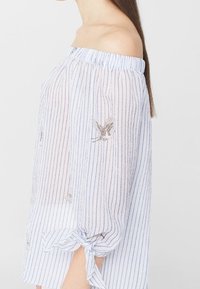 Off-Shoulder-Bluse aus hellblauem und weiß gestreiften strukturierten Stoff, mit bestickten Vögeln und gebundenen Ärmeln.
