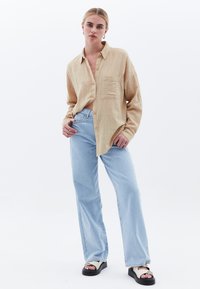 Camicia button-up beige con texture e una tasca, abbinata a jeans a gamba larga blu chiaro e sandali con plateau neri con dettaglio a strap.