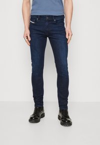Jeans de mezclilla azul oscuro con un corte ajustado, con un ligero desgaste, bolsillos frontales y un bolsillo para monedas. Llevados con botas negras con cordones.