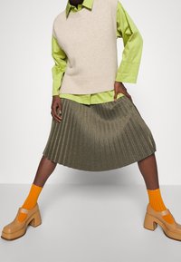 Maglione senza maniche beige su una camicia verde brillante, abbinato a una gonna plissettata oliva e calzini giallo-arancione con scarpe con zeppa marroni.