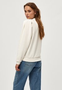 Sudadera blanca con costuras en los hombros abullonadas, puños y bajo de canalé, combinada con unos jeans azules holgados. Textura suave y corte relajado.