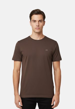 Giovane uomo che indossa una t-shirt marrone chiaro a maniche corte con un piccolo logo bianco sul lato sinistro del petto, in piedi davanti a uno sfondo bianco semplice.