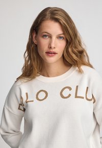 Weißer Strickpullover mit einem gerippten Rundhalsausschnitt und dem Text "BLO CLUB" in Braun auf der Vorderseite. Weiche Textur, lockere Passform.