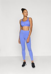 Μωβ αθλητικό σουτιέν με φερμουάρ, ταιριαστό ψηλόμεσο κολάν από απαλό ύφασμα. Το μοντέλο φοράει μαύρα αθλητικά παπούτσια, στέκεται μπροστά σε απλό φόντο.