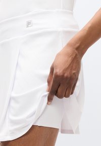 Λευκή αθλητική φούστα με εφαρμοστή σχεδίαση, διακριτικές κάθετες ρίγες και λογότυπο στη ζώνη μέσης. Ένα χέρι τραβάει το στρίφωμα.