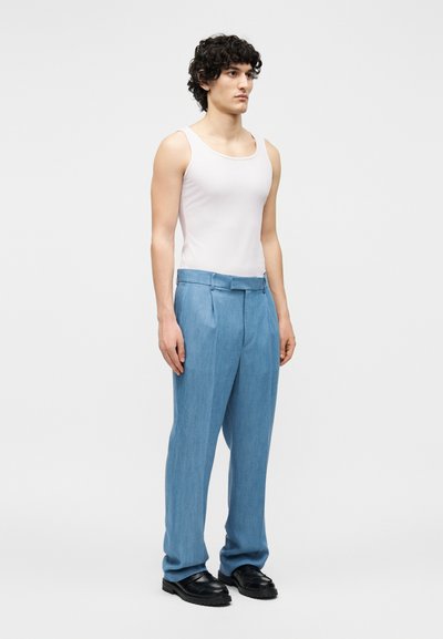 Jeune homme debout, portant un haut sans manches blanc, un pantalon bleu clair sur mesure et des chaussures en cuir noir sur un fond uni.