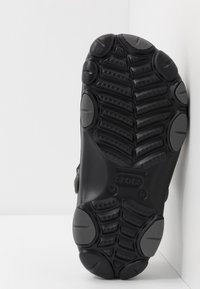 Semelle de chaussure en caoutchouc noir avec des motifs de grip texturés, présentant des bords arrondis et des détails d'accent circulaires en gris.