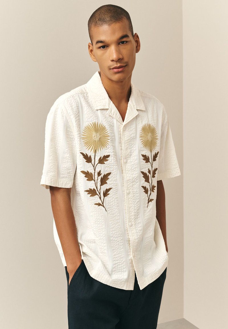 Giovane uomo che indossa una camicia bianca a maniche corte con due grandi girasoli ricamati, in piedi con le mani nelle tasche dei pantaloni neri.