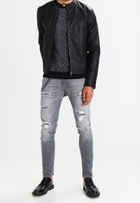 Schwarze Lederbomberjacke mit strukturierten Schultern, kombiniert mit einem strukturierten schwarzen Pullover und abgewetzten grauen Jeans. Schwarze Schuhe vervollständigen den Look.