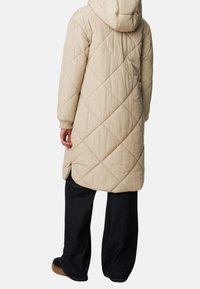 Abrigo largo acolchado en color beige con capucha, que presenta un diseño de costura en forma de diamante y puños acanalados, combinado con pantalones oscuros de corte holgado.