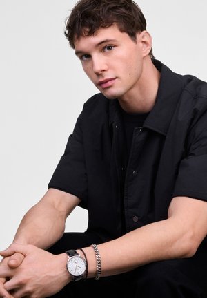 Chemise noire à boutons, à manches courtes, portée par-dessus une chemise noire. Montre bracelets noire avec détails argentés et bracelet en chaîne au poignet.