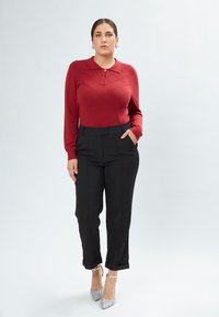 Camicia rossa aderente a maniche lunghe con motivo a rombi, abbinata a pantaloni neri a vita alta. Il modello indossa tacchi argentati glitterati e si posa con le mani in tasca.