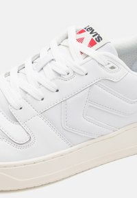 White synthetic sneakers with textured surface, featuring white laces, a cushioned sole, and a red and black logo tag on the tongue.