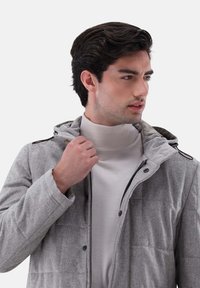 Grau isolierte Jacke mit Kapuze, mit einem gesteppten Design, schwarzem Zubehör und einem Reißverschluss vorne, getragen über einem hellen Rollkragenpullover.