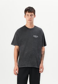Young man with short brown hair wears a faded black Nike SB t-shirt and black pants, standing against a plain white background.