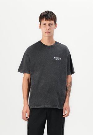 Un jeune homme aux cheveux bruns courts porte un t-shirt noir Nike SB délavé et un pantalon noir, se tenant devant un fond blanc uni.