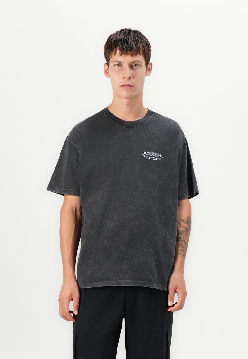 Young man with short brown hair wears a faded black Nike SB t-shirt and black pants, standing against a plain white background.