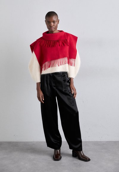 Pull tricoté rouge et blanc avec franges, associé à un pantalon large en satin noir et des bottes à talons épais, sur un fond neutre.