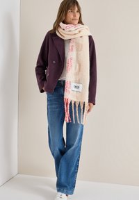 Strikket uldscarf i blød fersken med røde mønstre og frynsede kanter; bæres med en lilla jakke og blå vide jeans.