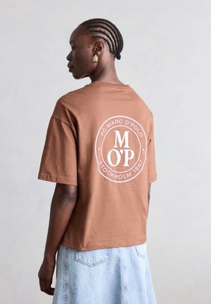 Γυναίκα με πλεξούδες, φορώντας καφέ μπλουζάκι με λευκό κυκλικό λογότυπο και ανοιχτή τζιν φούστα, σε πλαϊνή όψη με φόντο μονόχρωμο.