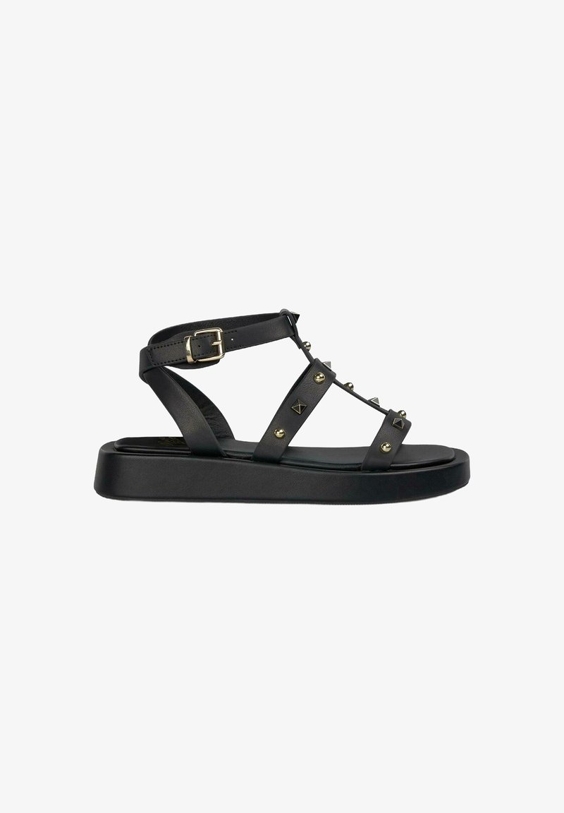 Zwarte platte sandalen met een brede zool, voorzien van een verstelbare enkelriem, metalen studs en decoratieve vormen op de riemen. Glad leren textuur.