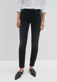 Jean skinny en denim noir avec une taille mi-haute, cinq poches, et un léger délavage. Associé à des chaussures noires sans lacets et une chemise blanche à boutons.