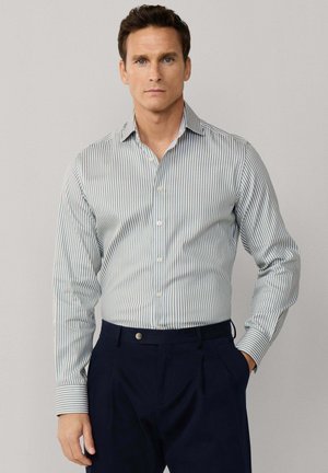 Homme portant une chemise à boutons rayée verticale bleu clair et blanc et un pantalon bleu marine foncé, debout devant un fond uni.