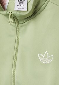 Světl zelená zipová bunda Adidas s texturovanou tkaninou, na levé straně s bílým logem a odpovídajícím jezdcem na zipu.