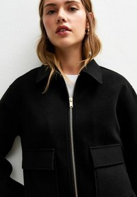 Veste en polaire noire avec un col, fermeture éclair à l'avant et deux poches plaquées. Texture lisse avec un design épuré et minimal.