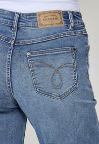Jeans de mezclilla azul claro con un diseño de bolsillo curvado y costuras en contraste color marrón. La etiqueta de la marca en la cinturilla dice "Koroshi."