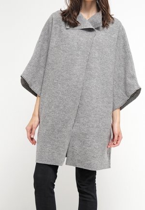 Frau trägt einen midilangen, grauen Wollmantel mit breiten Ärmeln über schwarzen Hosen, stehend vor einem schlichten weißen Hintergrund.