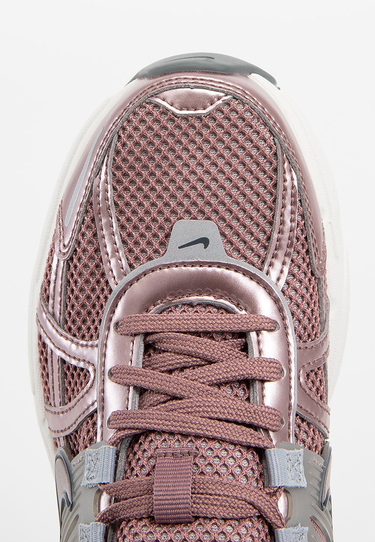 Zapatilla Nike con parte superior de malla en oro rosa, acentos metálicos, lengüeta acolchada y detalles en gris. Presenta cordones texturizados y una suela blanca.