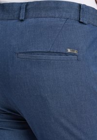 Dunkelblaue Denim-Hosen mit einer Gesäßtasche mit einem Metall-Logo-Akzent und einem flachen Bund. Der Stoff hat eine glatte Textur.